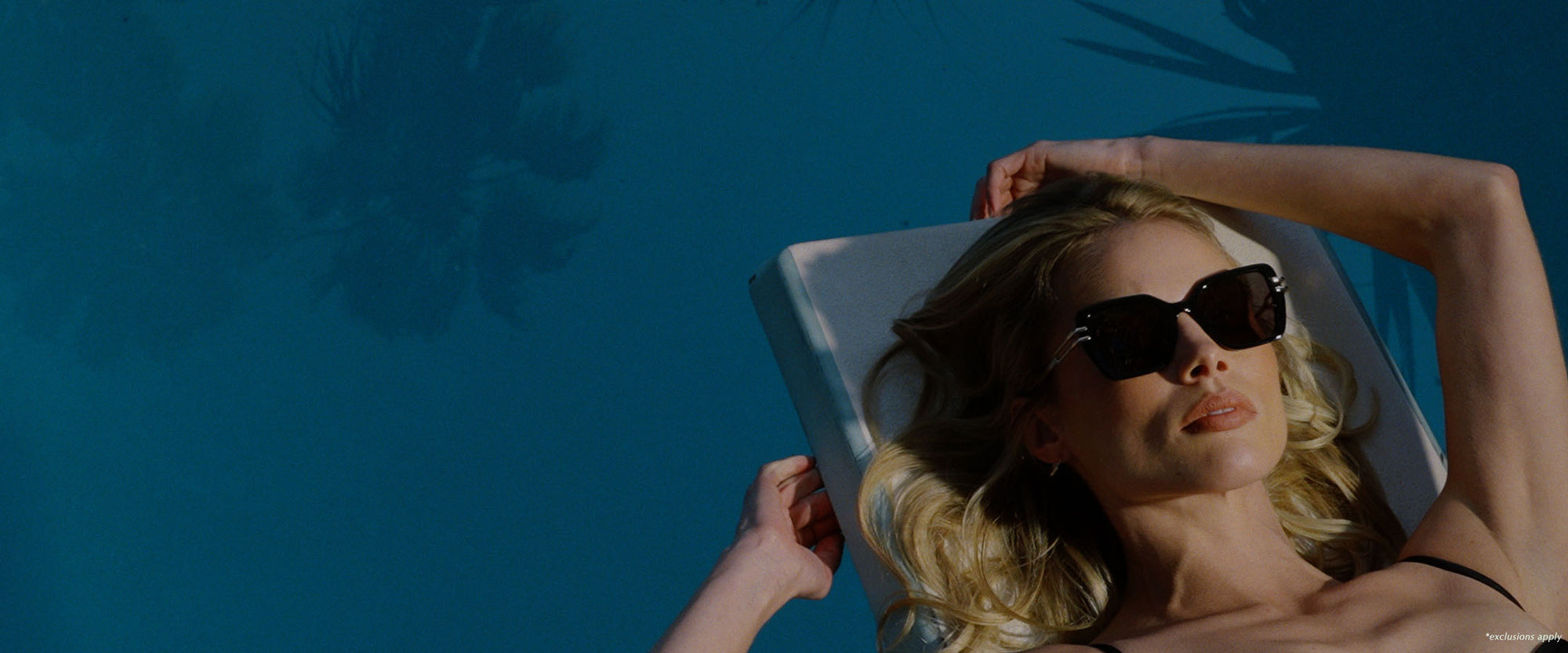 Blonde woman with black one piece and black oversized sunglasses poolside