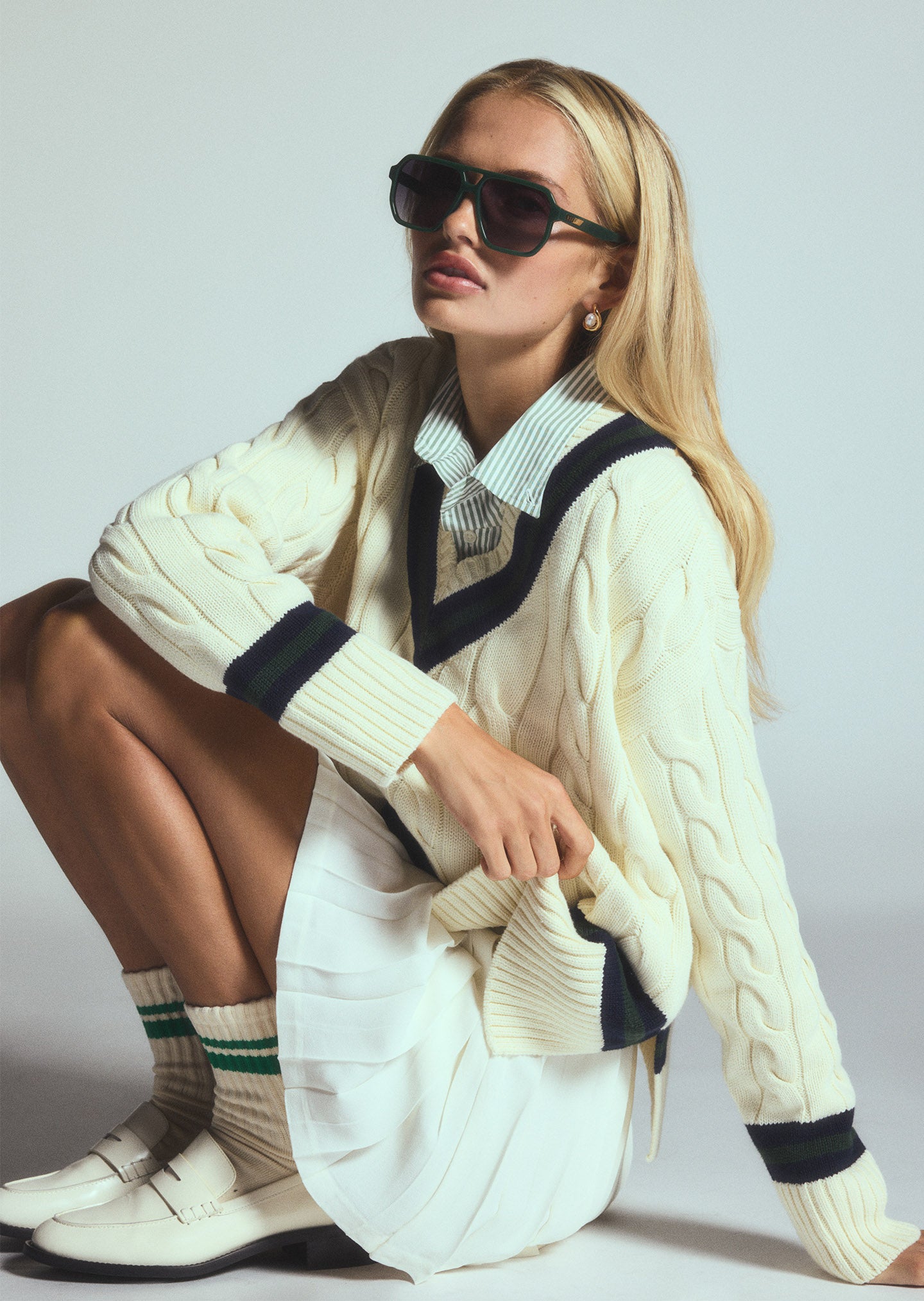 Color studio shot of blonde woman wearing a white cable-knit cricket sweater and white mini skirt kneeling down while wearing Cameron sunglasses
