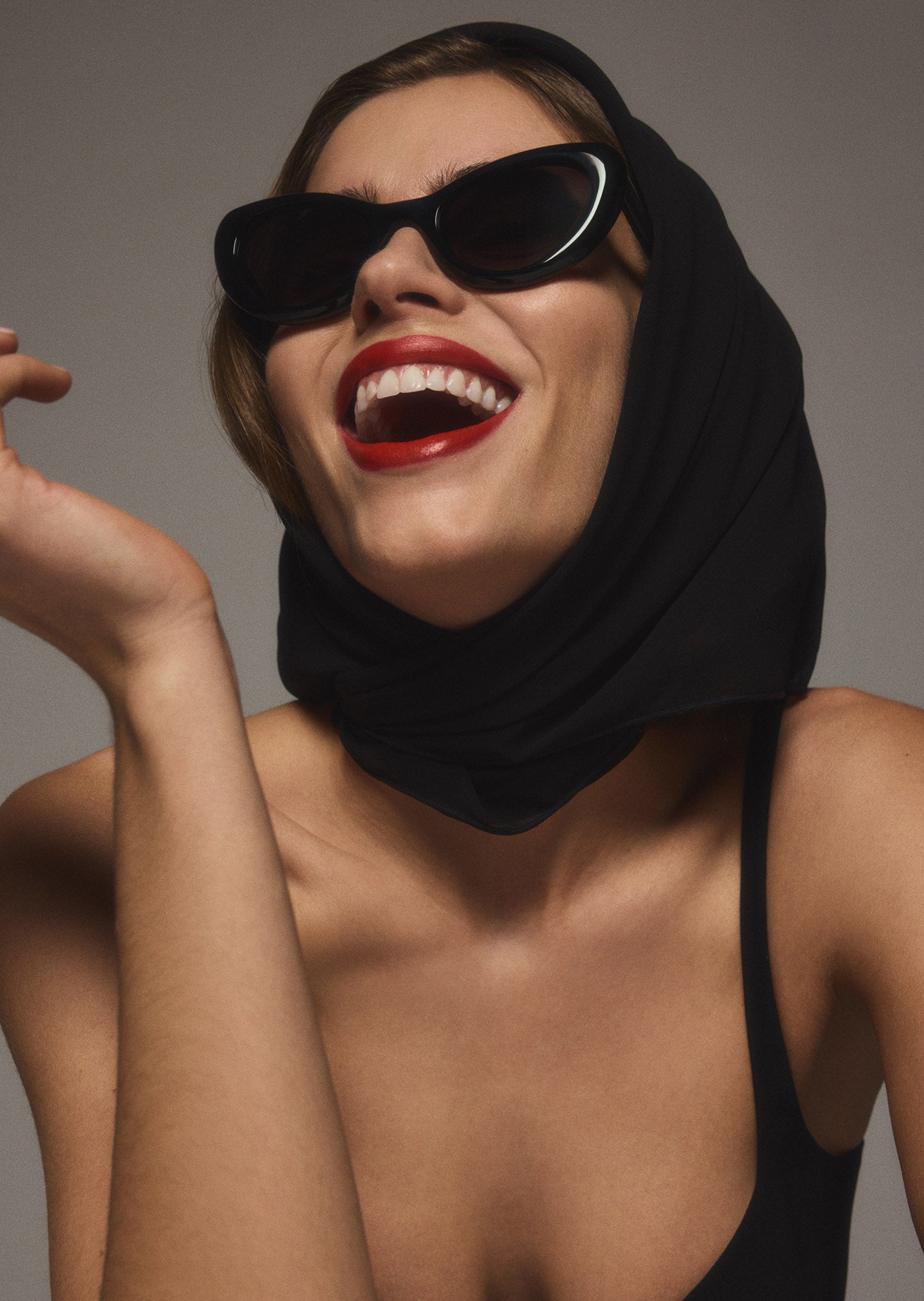 Studio shot of brunette haired woman with red lipstick glancing upward with a large smile while wearing black Zaria sunglasses