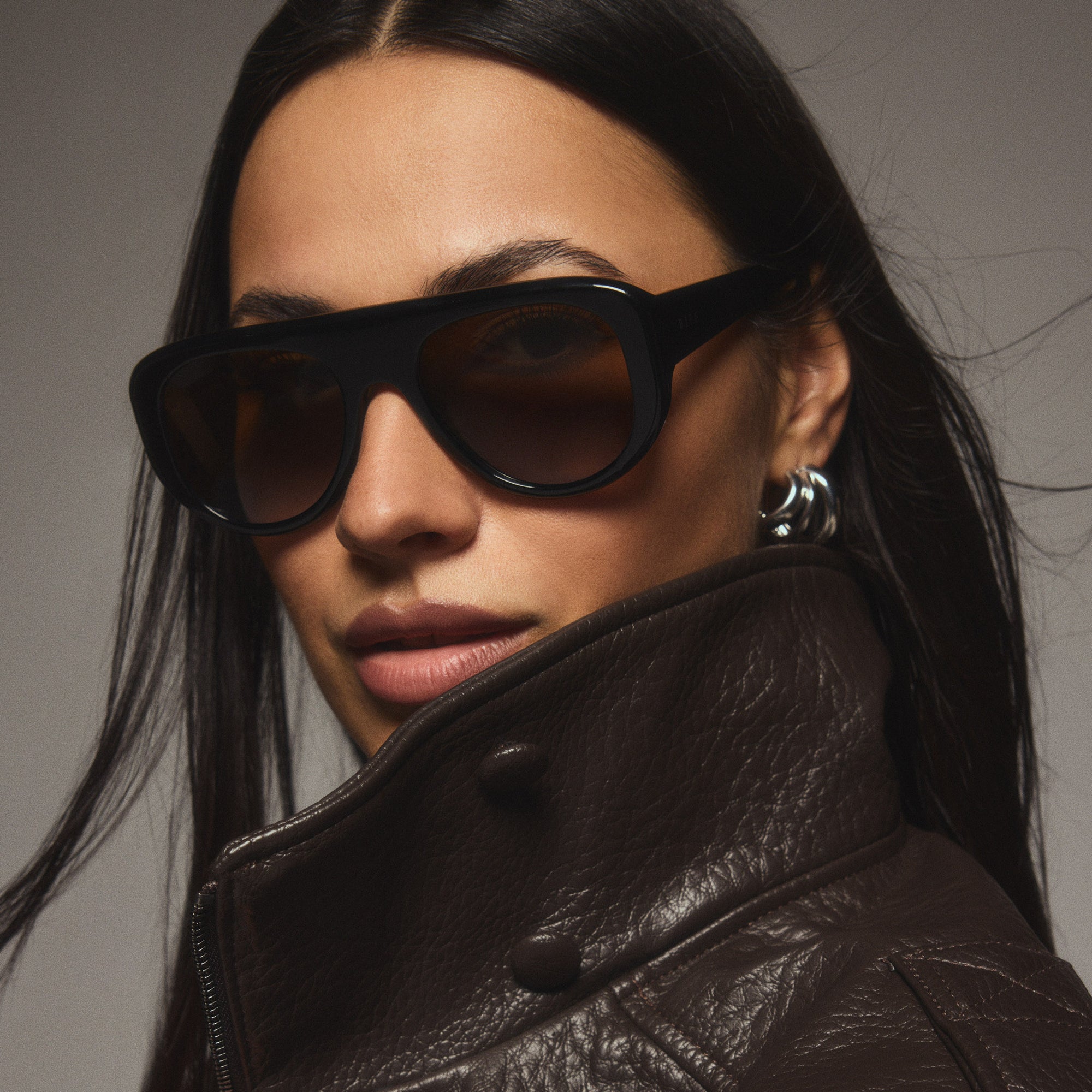 Studio shot of dark-haired woman wearing thick dark brown leather jacket glancing over her left shoulder at the camera while wearing black Morgan sunglasses