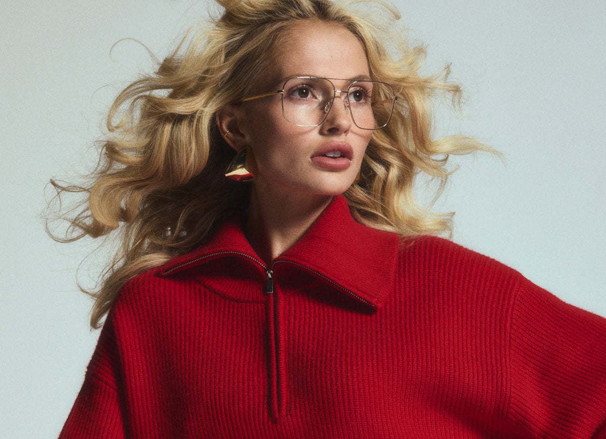 Studio shot of young blonde woman wearing stylish red quarter zip sweater looking off camera wearing Jonas gold eyeglasses