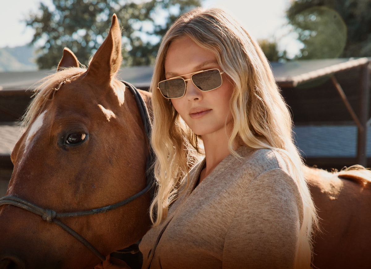 blonde woman wearing yellowstone pioneer aviator gold brown sunglasses in ranch with brown horse western fashion