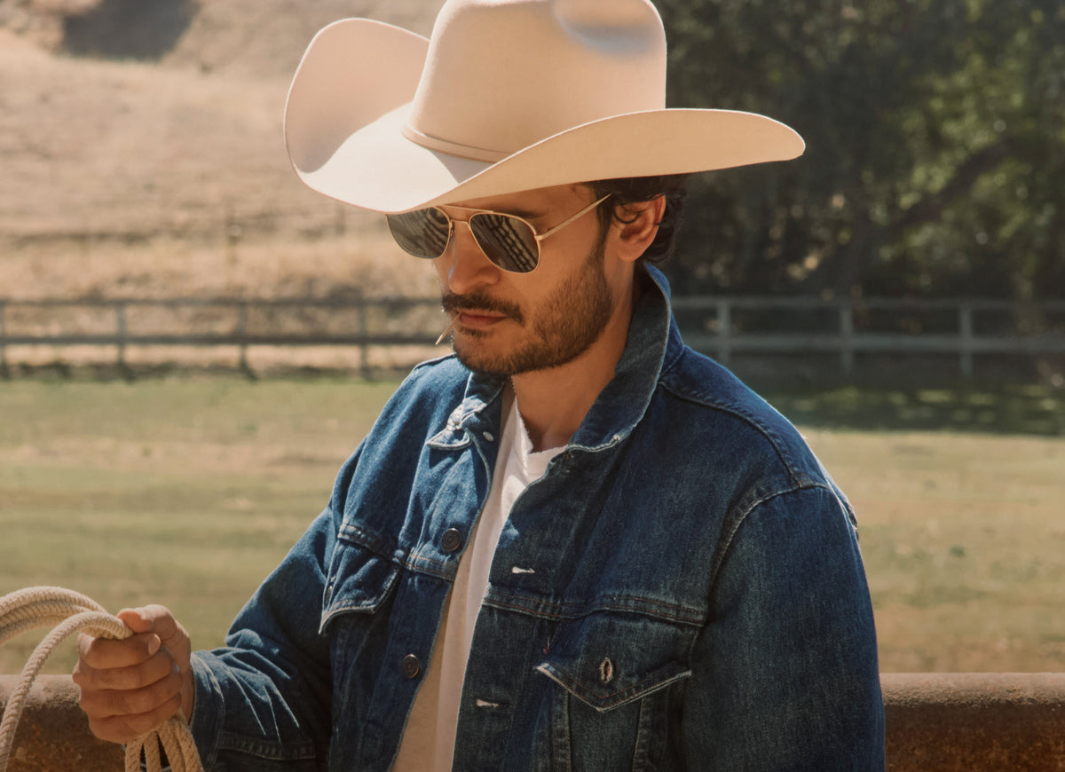 Man wearing cream colored cowboy hat in ranch wearing yellowstone the ranch sunglasses with denim jacket