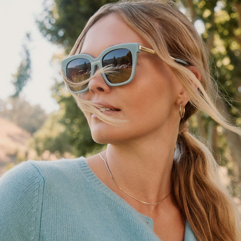 lifestyle - blonde female wearing diff eyewear bella square oversized sunglasses with a dew drop blue frame, gold temples and brown gradient lenses angled view